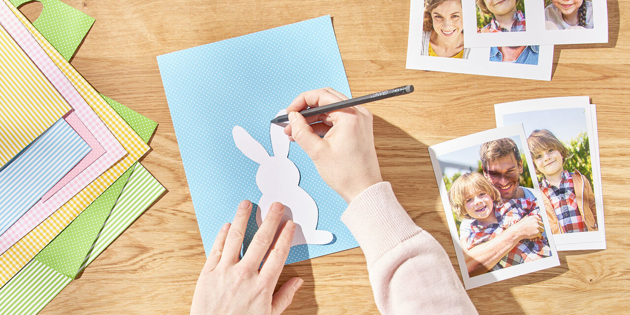 Két kéz rajzol egy húsvéti nyuszit a papírra a sablon segítségével. Mellette a család kinyomtatott azonnali fotói.